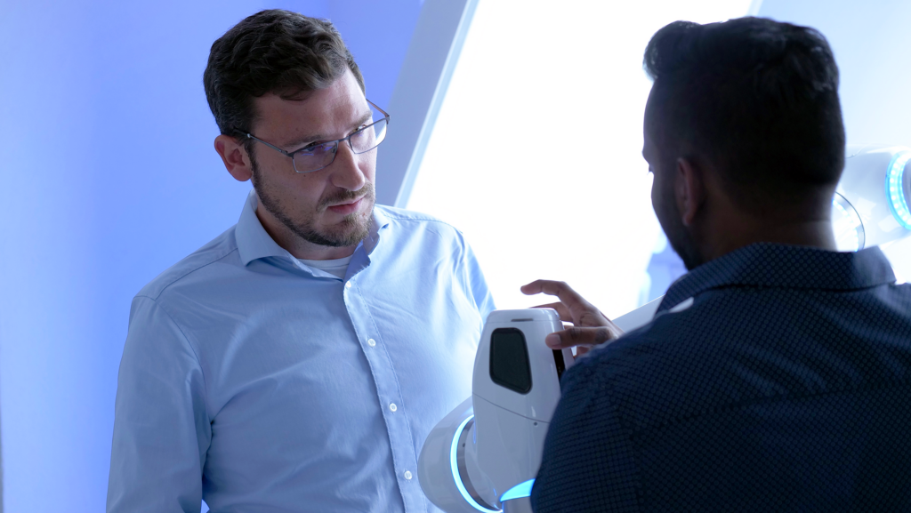 Arne Nordmann in conversation with a colleague beside a NEURA robot arm, from the start of his time at NEURA Robotics.